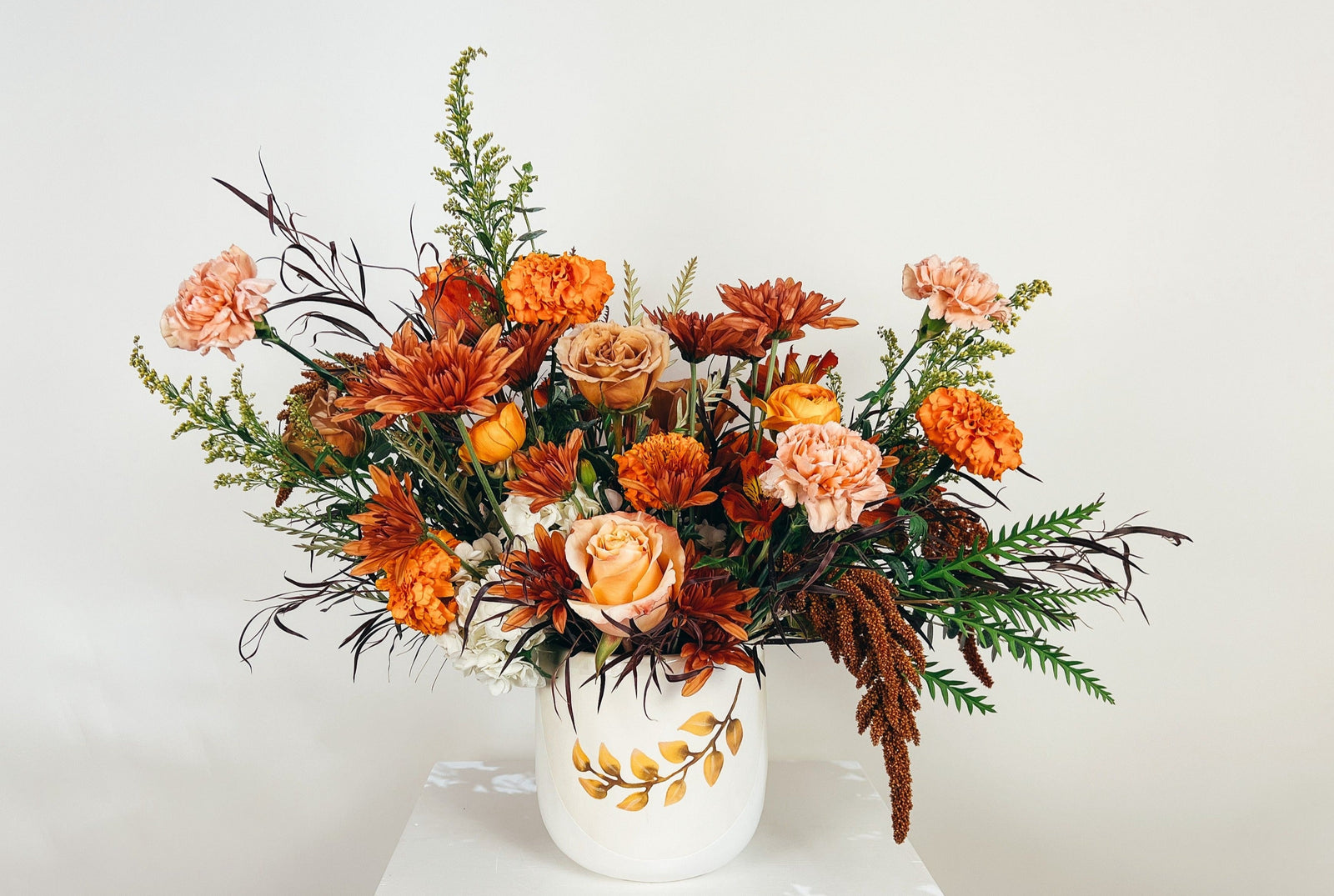 A floral arrangement featuring a ceramic pot with a harvest leaf emblem, containing a mix of peach, orange, and bronze colored flowers such as roses and mums, with green foliage and bronze amaranthus.
