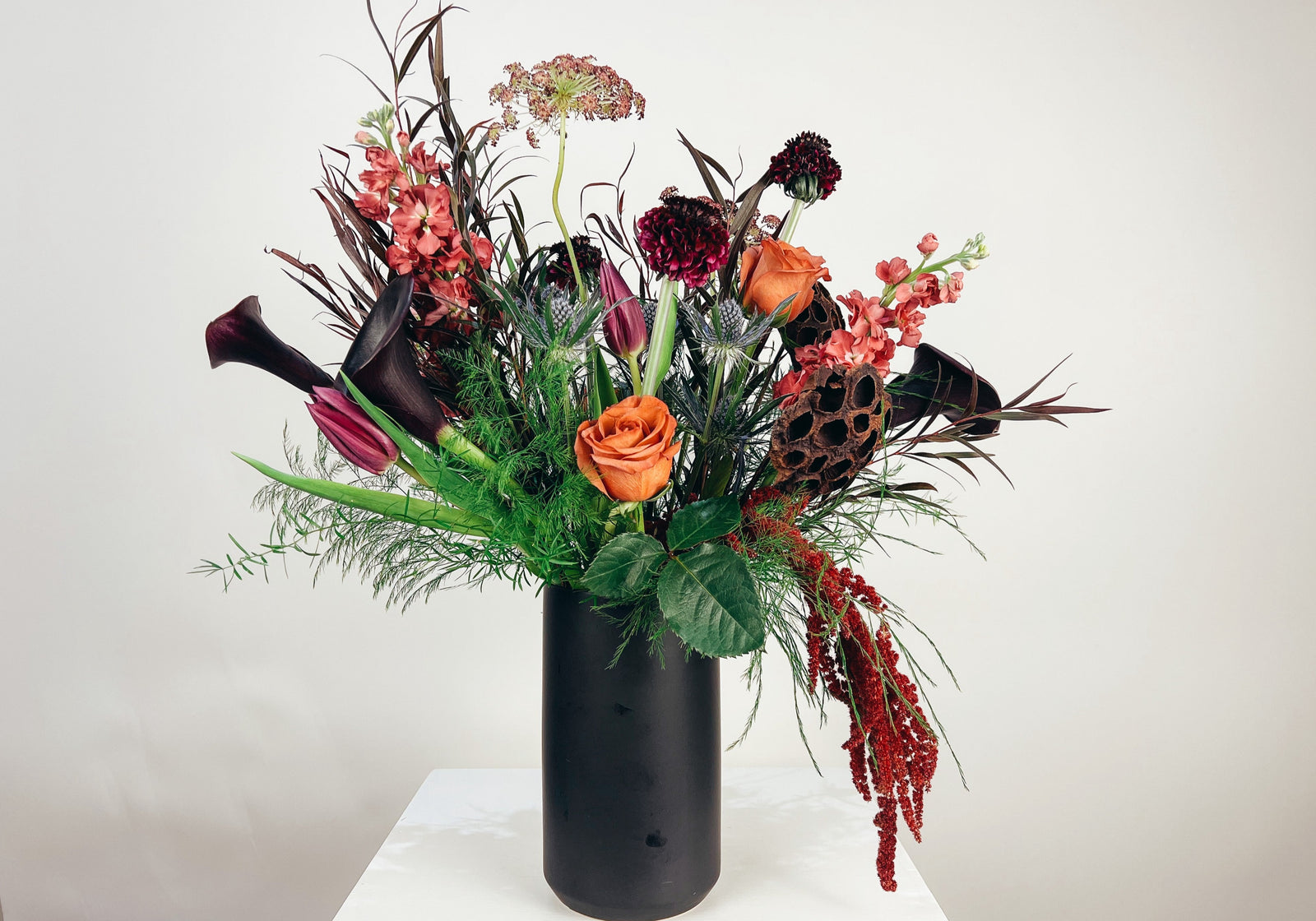 A Halloween-themed floral arrangement featuring a variety of flowers including black Calla Lilies, deep purple Scabiosa, Chocolate Queen Annes Lace, and more, in shades of black, purple, orange, and green, delivered in a matte black ceramic vase.